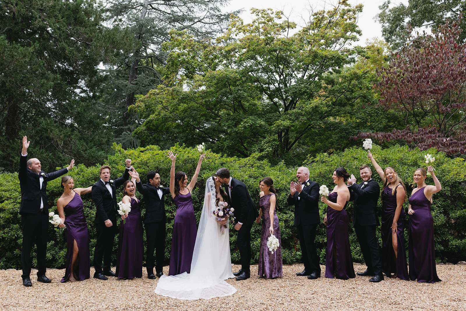 A wedding party in deep purple and black tie cheering with raised bouquets while the couple kiss on a gravel path at VanLandingham Estate